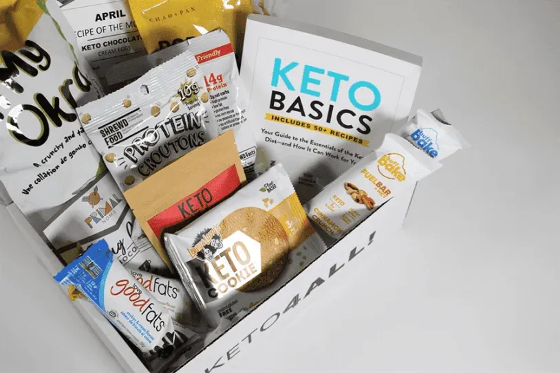 A Keto Basics box filled with various keto-friendly snacks including protein chips, cookies, and other packaged goods. Main text on image: "KETO BASICS," "INCLUDES 8+ RECIPES".