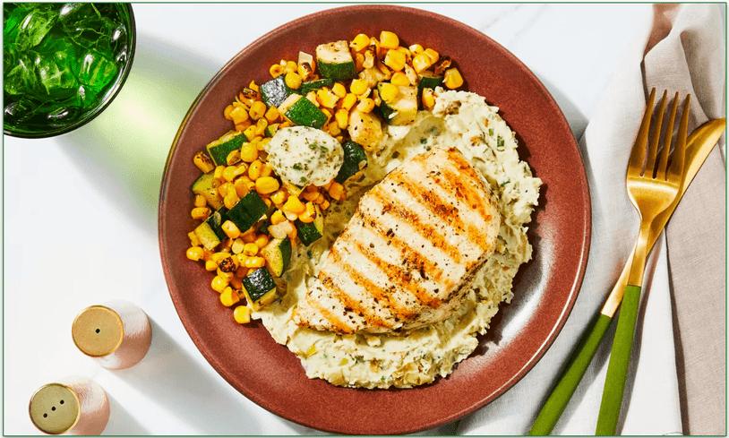 A plate of chicken atop mashed potatoes with a side of carrot and zucchini