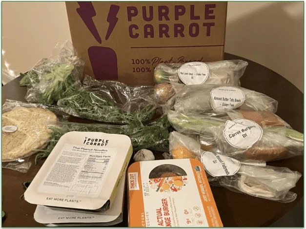 Photo of 4 recipe ingredient bags, prepared meals and other ingredients in front of a Purple Carrot delivery box