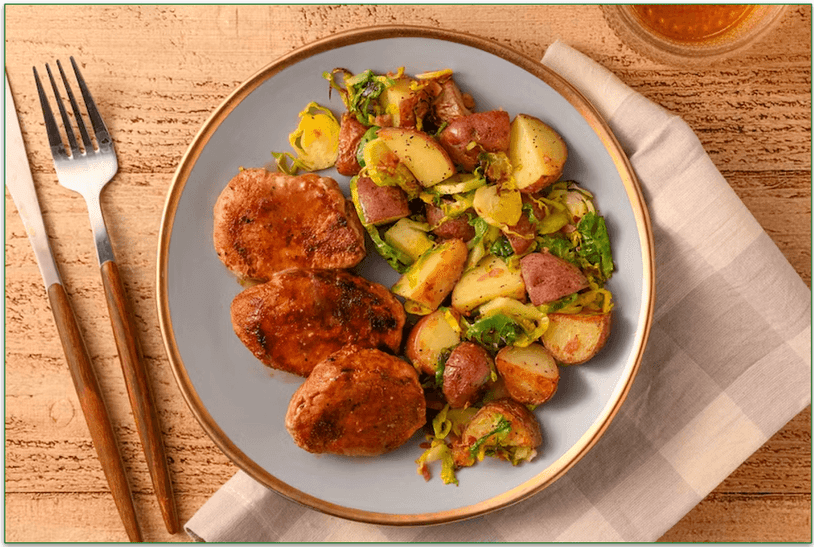 A plate of pork tenderloin beside a side order of roasted  Brussels sprouts and potatoes