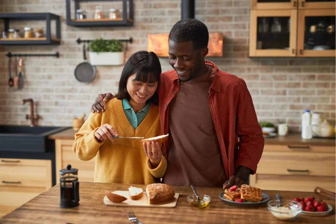A smiling couple in a kitchen spreads jam on bread. Wooden shelves and cooking utensils adorn the brick wall. The main text reads: "Happy Cooking Together."