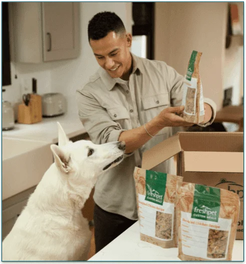 a man with his dog unpacking a Freshpet box on a kitchen counter