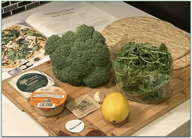 Photo of the ingredients for the Roasted Broccoli Flatbreads recipe on a board in front of the recipe in the booklet