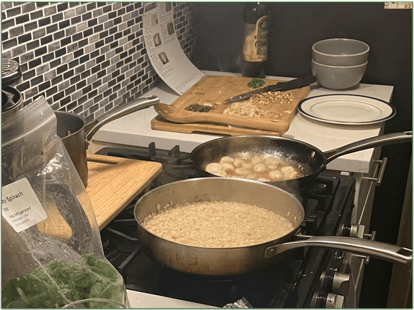 Two frying pans on a stove with a chopping board next to the stove and an open bag of greens