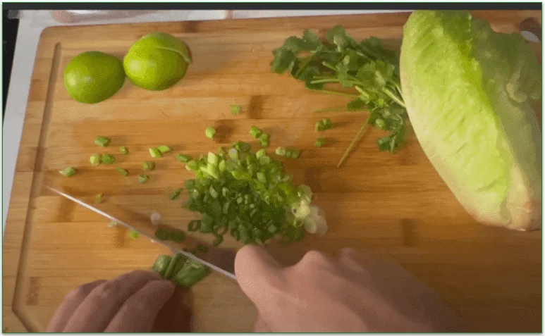 A board with limes, scallions, herbs, and lettuce being chopped