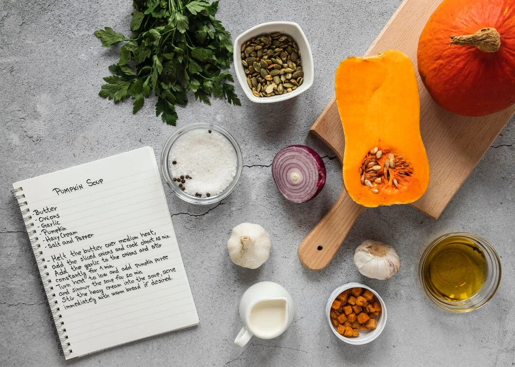 Ingredients for pumpkin soup are arranged on a gray surface: half a pumpkin, onion, garlic, parsley, pumpkin seeds, olive oil, cream, salt, pepper, and turmeric. A notebook with a handwritten recipe for "Pumpkin Soup" is also visible.