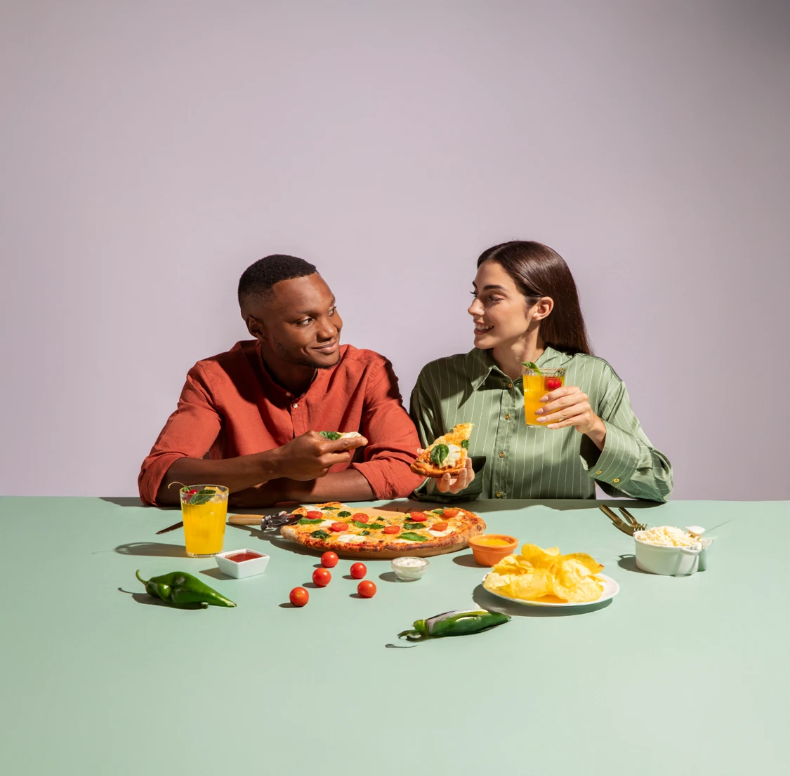 A man and woman sit at a table with pizza, chips, and drinks. They are smiling and sharing slices. Text on image: "Delicious Moments."
