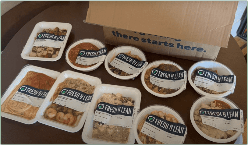 Photo shows 10 meals in trays and bowls displayed on a table next to a Fresh N Lean delivery box