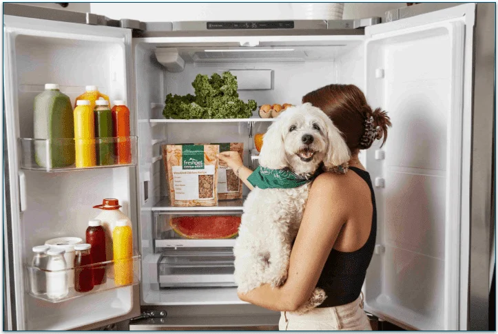 a woman holding a small white fluffy dog reaching into the fridge for  a Freshpet pack