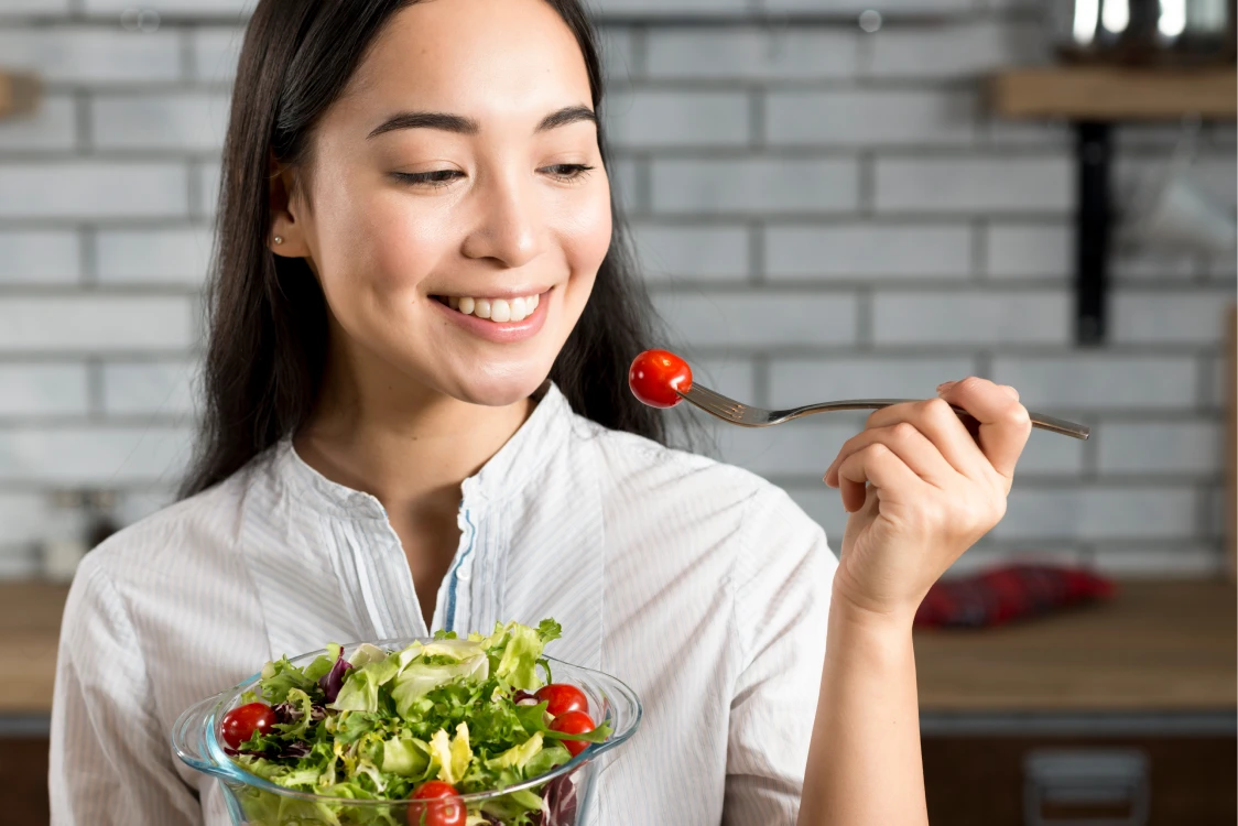 A woman smiles while holding a fork with a cherry tomato. She also holds a bowl of mixed salad with leafy greens and tomatoes. The background features a brick wall and kitchen shelves.