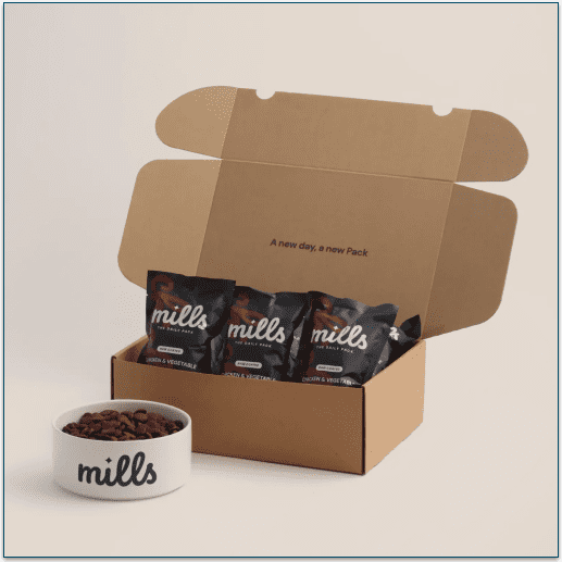 Picture of an open cardboard box containing Mills Daily Packs next to a dog food bowl
