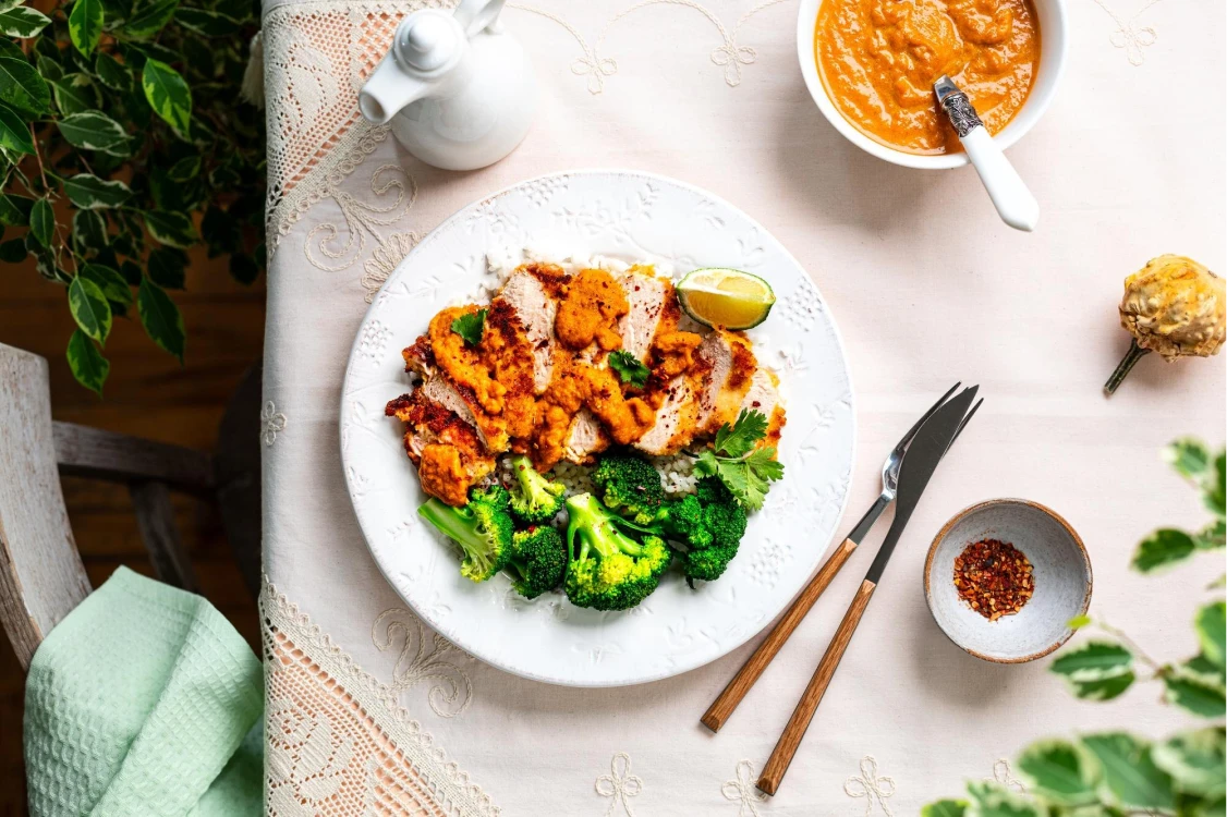 A white plate with grilled chicken, broccoli, and a slice of lime, topped with orange sauce. A small bowl with more sauce and a spoon is nearby. The text on the image says "Simple Dinner Recipe." A fork, knife, and small dish of chili flakes are on the table.