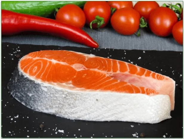 A fish steak on a cutting board beside a hot pepper, tomatoes, and a cucumber.
