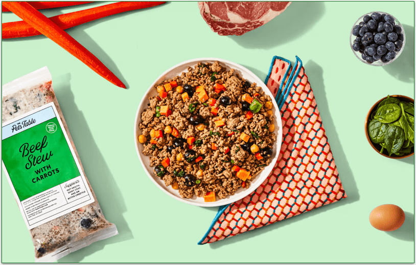 Image of The Pets Table's Beef Stew fresh recipe in a bowl surrounded with fresh ingredients and a pack of the recipe