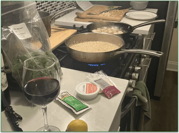 Photo of 2 dishes cooking on the stove (risotto and scallops), chopped veg on a chopping board in the background, and a glass of red wine and other ingredients in the foreground