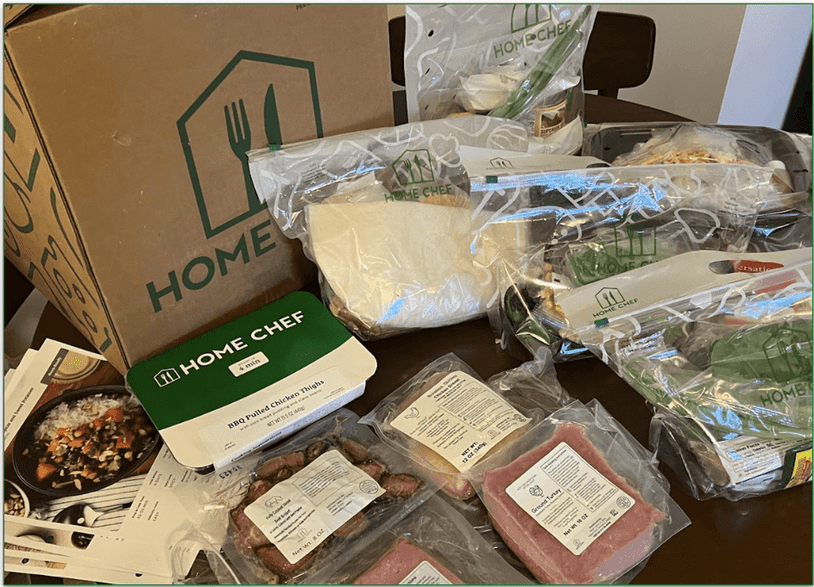 Selection of Home Chef branded meal kits and a prepared meal on a table next to a Home Chef delivery box