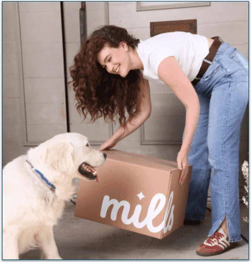 A woman picking up a Mills cardboard box and a golden retriever watching