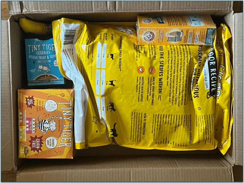 Image of an open cardboard box containing a large bag of dry cat food, a box of canned cat food, a box of cat treats, a toy, and a box of cat litter deodorizer