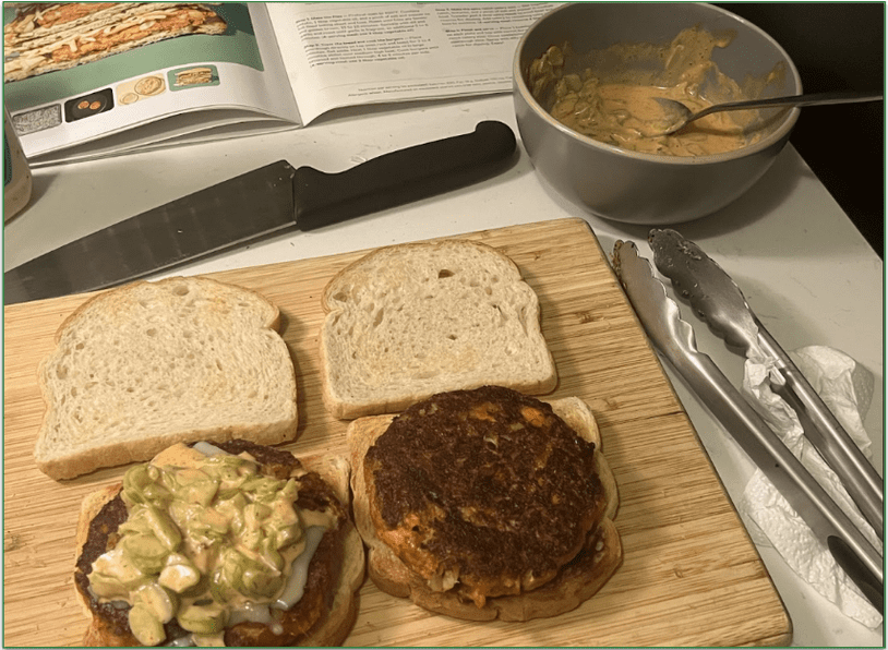 Photo of the assembly stage of the Carrot Burgers recipe with the burgers on a wooden board next to the open recipe and a bowl of dressing